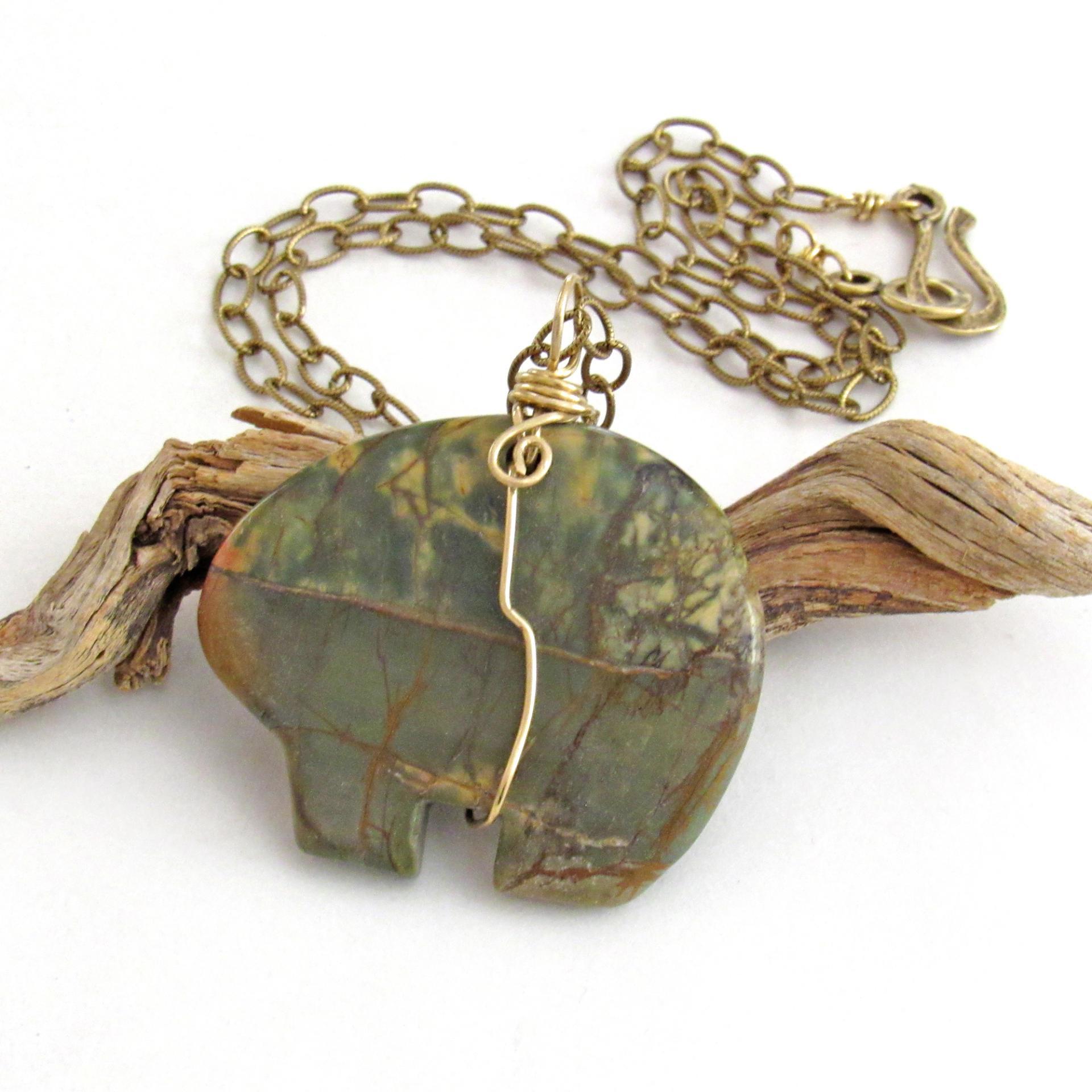 This pendant necklace features a large natural Red Creek jasper stone carved into a southwestern zuni bear shape. It's wrapped in gold brass wire and hangs from a textured brass chain. The stone has an earthy mossy green hue with subtle accents of yellow and streaks of brown. The style is unisex - perfect for a man or woman. One of a kind - it makes a great gift for a rock hound - or zuni bear lover.